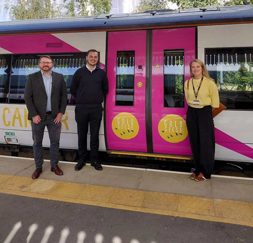 Two men in dark jackets and a woman in a yellow jumper stand on the platform beside the pink and white Gold Geese train, whose doors carry the charity's yellow circular logo