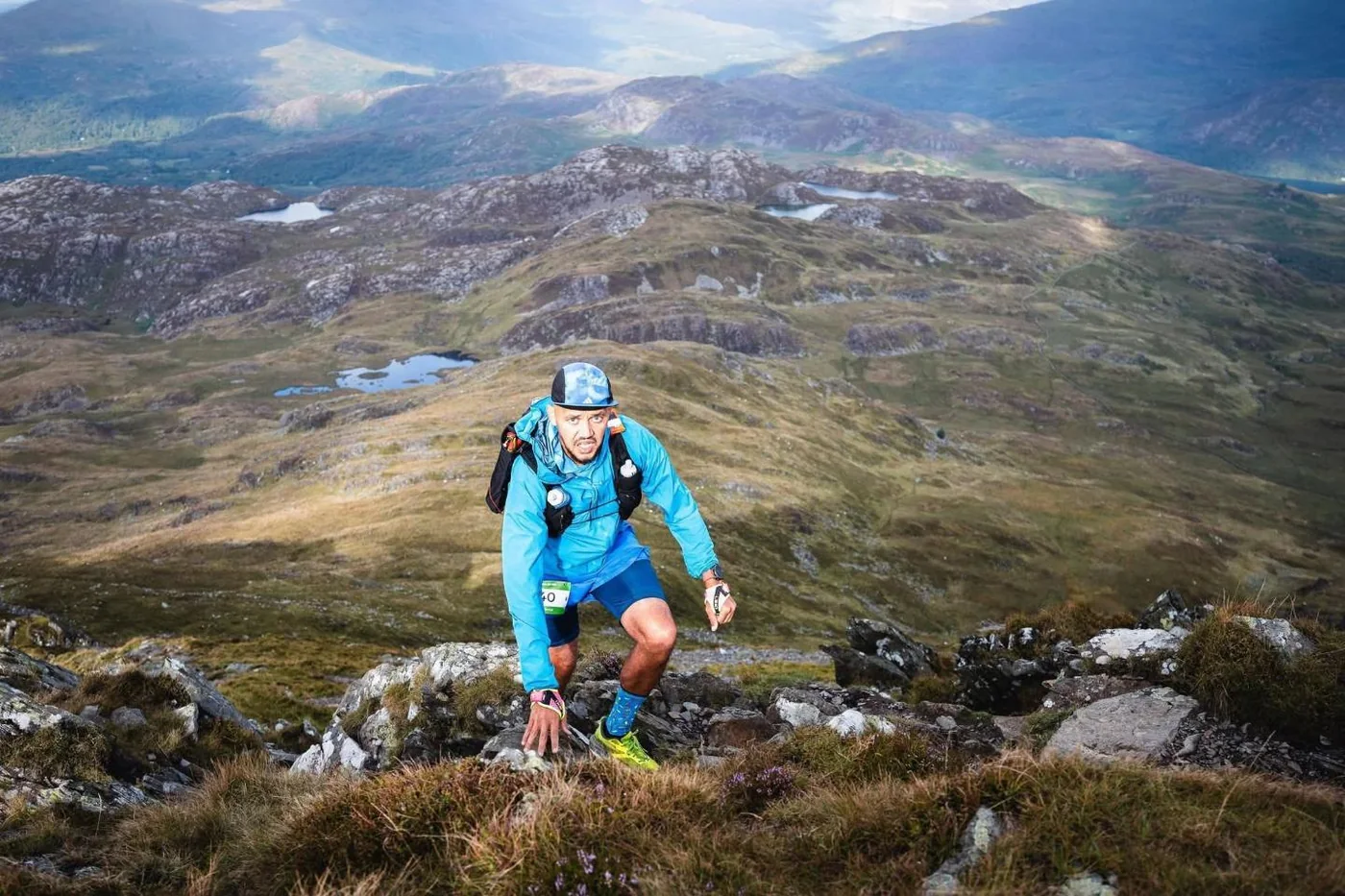 A runner using both hands on the rock to climb, with lakes on the plateau behind
