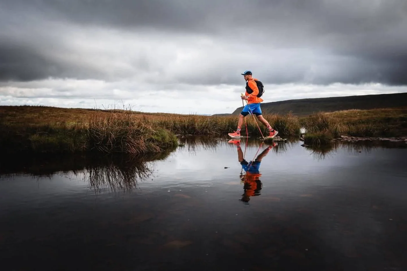 A runner crossing boggy moorland under heavy cloud, reflected in standing water