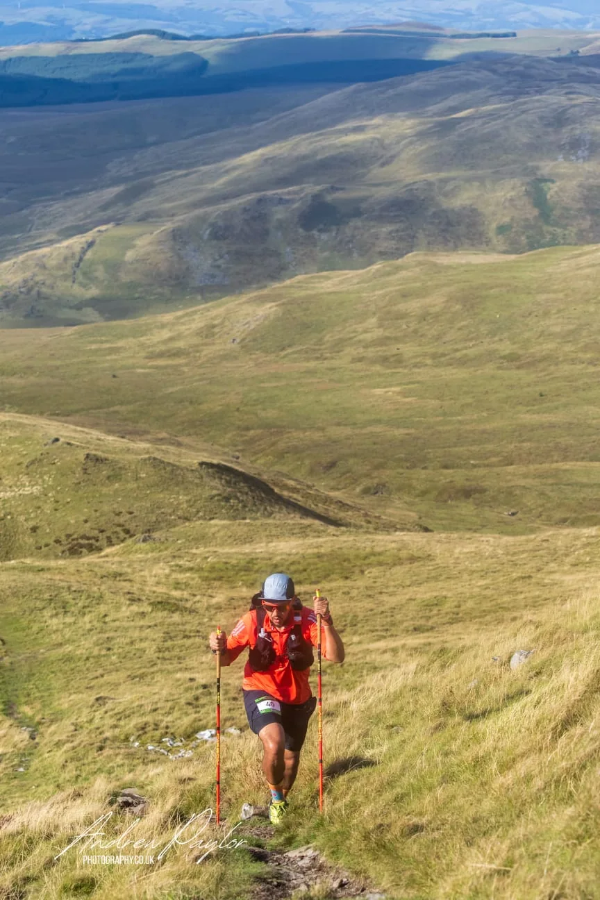 A runner using poles on a steep grass slope, with a wide Welsh valley behind