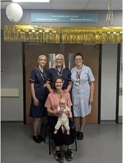 Four members of Southend Hospital's maternity and infant feeding staff in navy and blue uniforms stand beneath gold tinsel outside the Fetal Medicine Centre, with a seated mother holding a baby in front of them